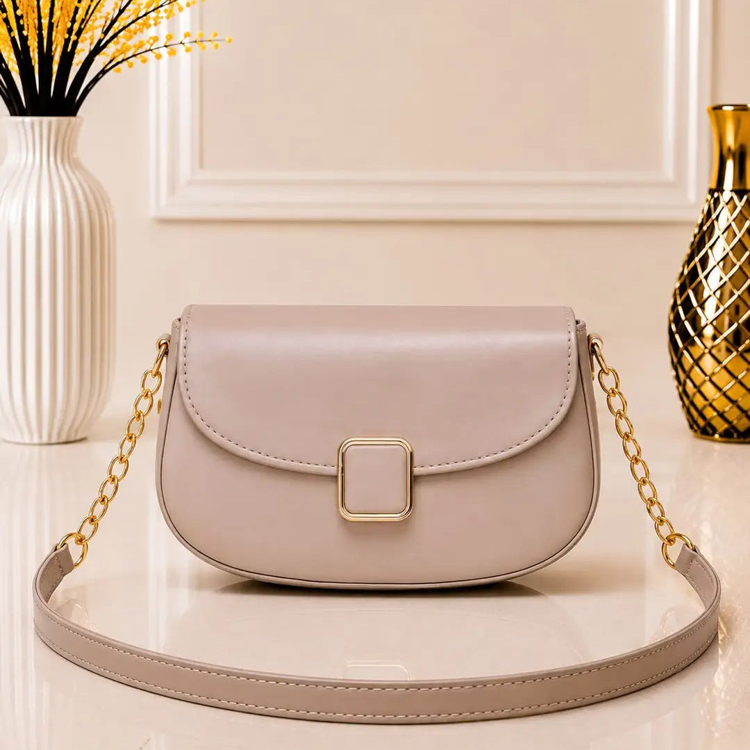 A light beige leather shoulder bag with a gold chain strap and a square gold buckle, displayed on a glossy white surface with decorative vases in the background.